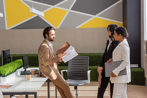 side view of young manager with documents talking to multiethnic businesswomen with laptop and folder in modern office