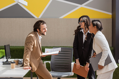 smiling businessman holding documents and talking to multiethnic colleagues with laptop and folder in office