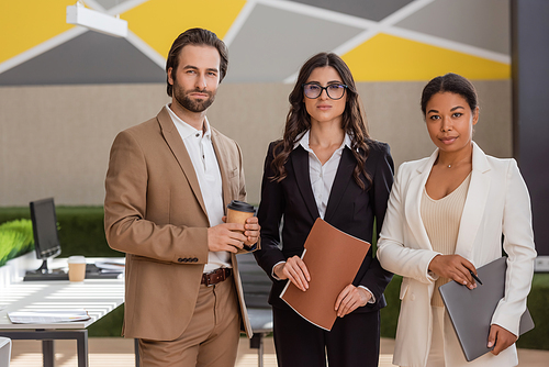 multiethnic business people with laptop and coffee to go looking at camera in office