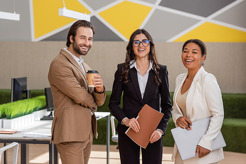 cheerful multicultural managers in formal wear smiling at camera in modern office