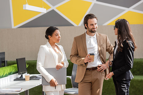 smiling manager standing with hand on hip and coffee to go near interracial businesswomen in office