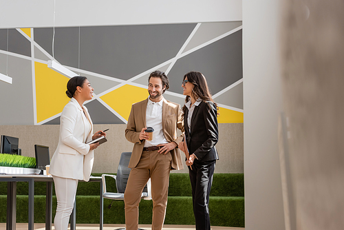 happy multiethnic managers in formal wear talking near desk with computers in modern office