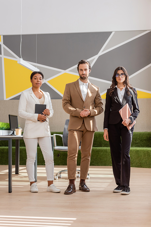 full length of multicultural business people looking at camera while standing in modern office