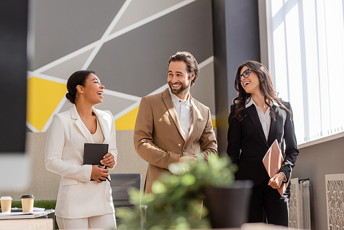 multiethnic business people in formal wear talking and laughing in office on blurred foreground