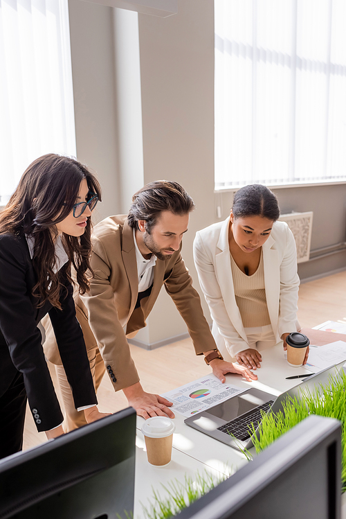 multiethnic business colleagues looking at laptop near charts and paper cups on desk in office