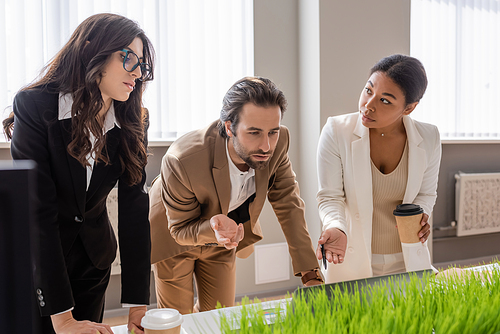 young manager and multiracial businesswoman with coffee to go pointing at laptop near colleague in eyeglasses in office