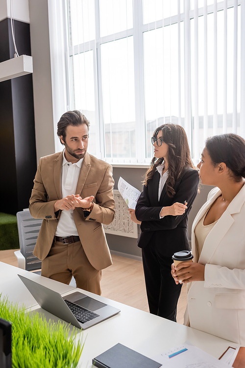 young manager counting on fingers while talking to multiethnic businesswomen near laptop on desk in office