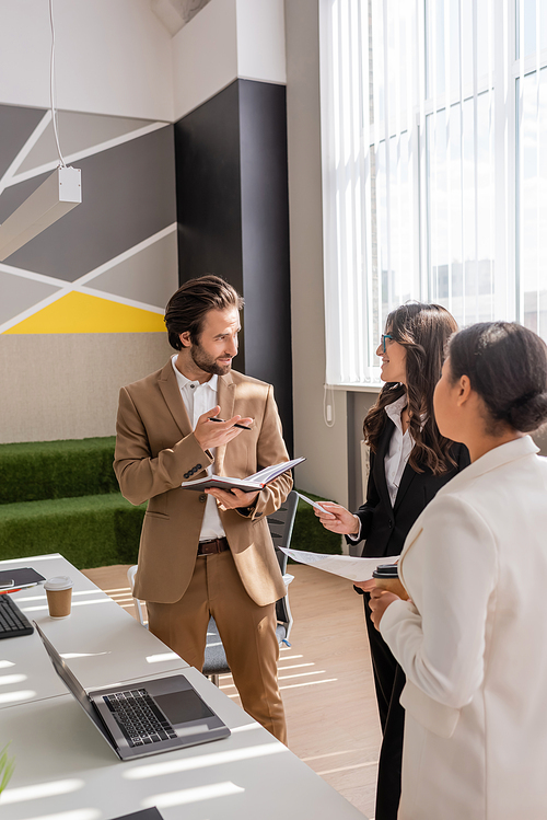 young manager with notebook talking to multiethnic colleagues near laptop on desk in office