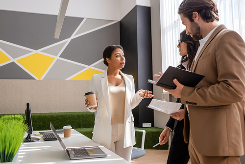 multiracial businesswoman with paper cup talking to colleagues near desk with computers in office