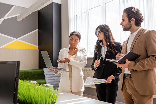 multiracial businesswoman pointing at laptop near colleagues with notebook and graphs in office