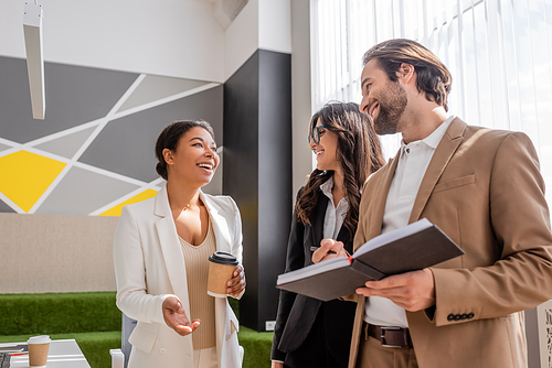 cheerful multiracial businesswoman with paper cup talking to smiling managers in office