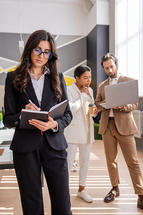 businesswoman in eyeglasses writing in notebook near thoughtful interracial colleagues looking at laptop on blurred background