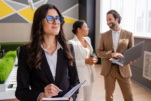 smiling businesswoman in eyeglasses holding notebook and looking away near blurred colleagues with laptop and takeaway drink