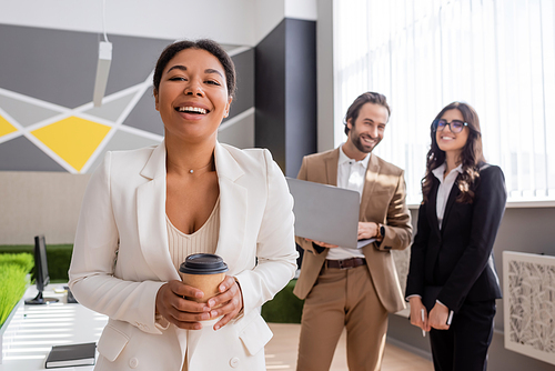 cheerful multiracial businesswoman with coffee to go looking at camera near colleagues with laptop on blurred background