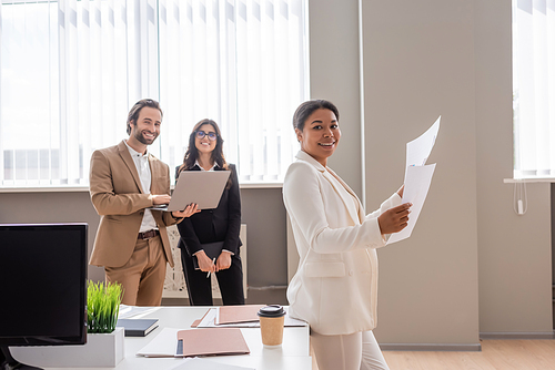 multiracial businesswoman holding papers near managers with laptop smiling at camera in office