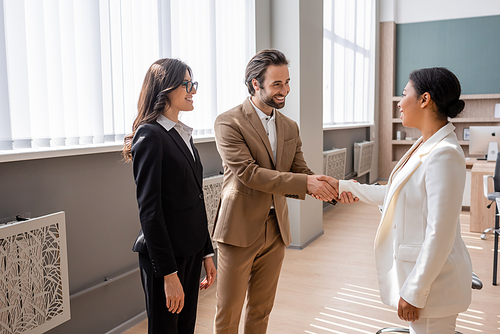 young and happy manager shaking hands with multiracial businesswoman near colleague in office