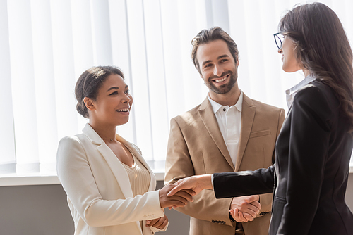 multiethnic businesswomen in formal wear shaking hands near young and smiling manager in office