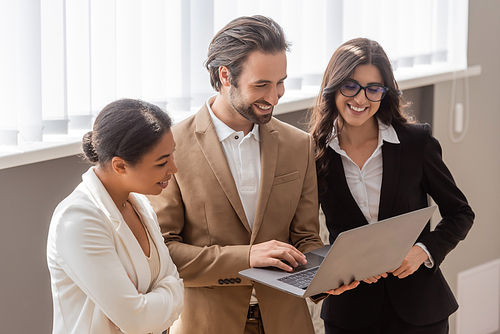 happy businessman using laptop near multiethnic colleagues standing in office