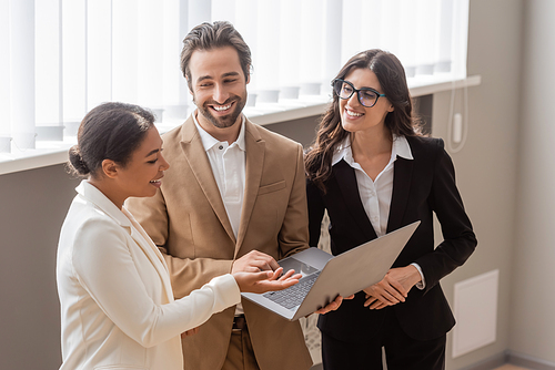 multiracial businesswoman pointing at laptop near cheerful managers in office