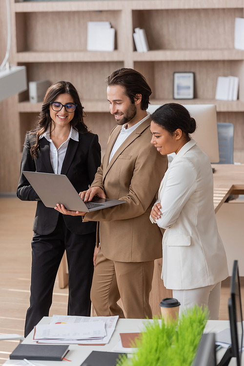 smiling manager showing laptop to interracial businesswomen near documents and paper cup on desk in office