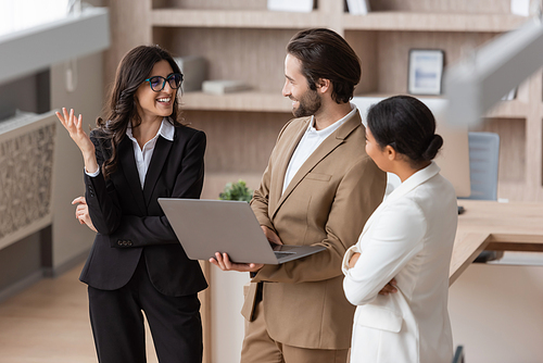 multiracial businesswoman and man with laptop looking at cheerful colleague in eyeglasses talking and gesturing in office