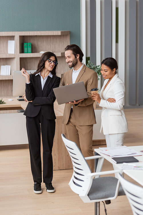 cheerful manager standing with laptop near interracial businesswomen in modern office