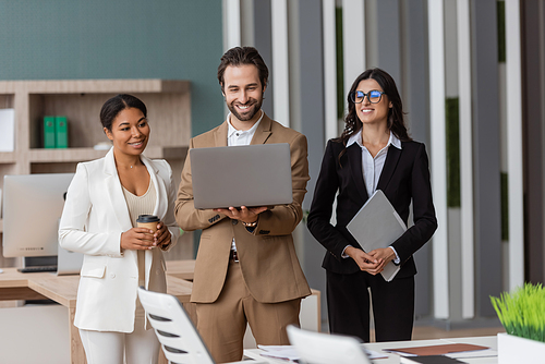 young and happy manager using laptop near interracial businesswomen standing with folder and paper cup in office