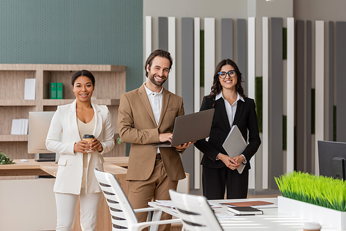 young manager with laptop and interracial businesswomen with paper cup and folder smiling at camera in office