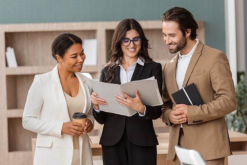 smiling manager in eyeglasses holding folder with documents near multiethnic colleagues with notebook and coffee to go