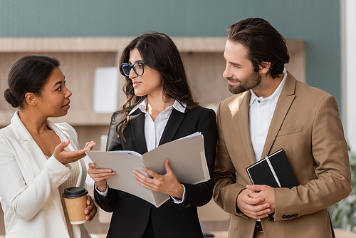 multiracial businesswoman with coffee to go talking to managers with folder and notebook in office