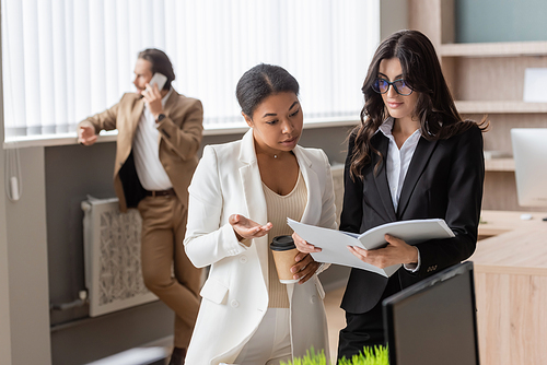 multiethnic businesswomen looking at documents near young manager talking on smartphone on blurred background