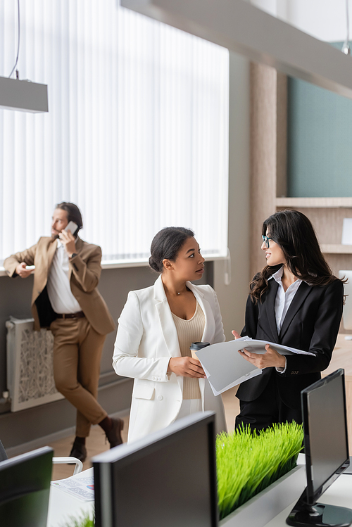 interracial businesswomen with paper cup and documents talking near blurred manager calling on smartphone in office