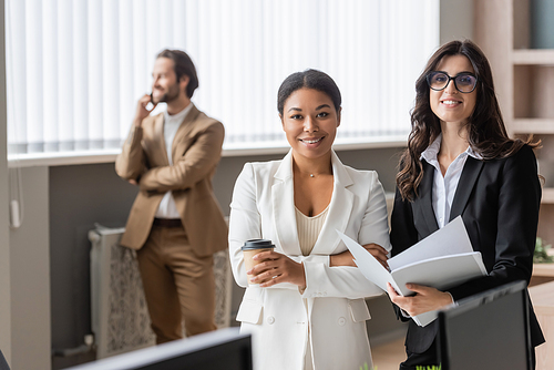 multiethnic businesswomen with paper cup and documents looking at camera near young manager on blurred background