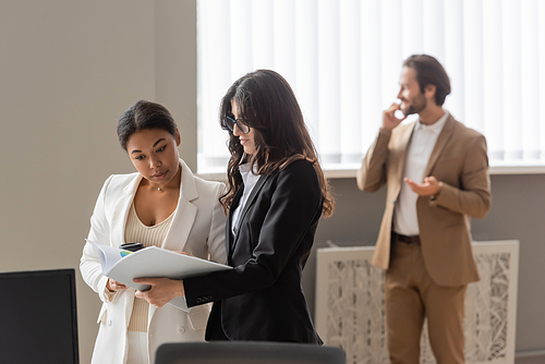 interracial businesswomen in formal wear looking at papers near young manager on blurred background