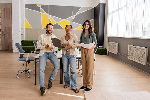 full length of happy multiethnic business people with gadgets and documents looking at camera in modern and spacious office