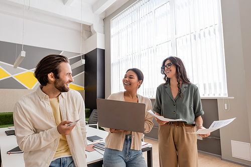 cheerful multicultural business people with gadgets and documents talking in modern office