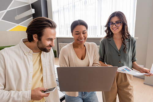 multiracial businesswoman holding laptop near colleagues with infographics and smartphone in office