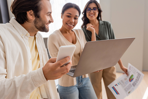 smiling manager holding smartphone near interracial businesswomen with laptop and documents on blurred background