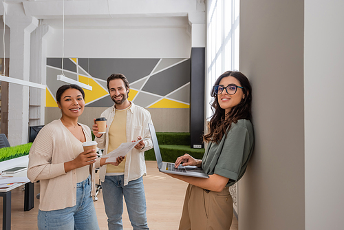 cheerful multiethnic business colleagues with gadgets and coffee to go looking at camera in office