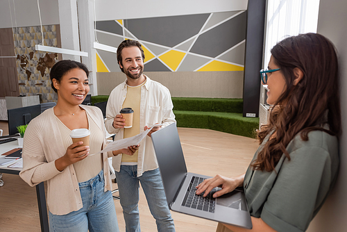 happy interracial managers with coffee to go looking at colleague standing with laptop in office