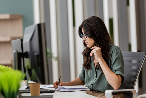 brunette manager in eyeglasses writing in notebook near computer monitors on blurred foreground