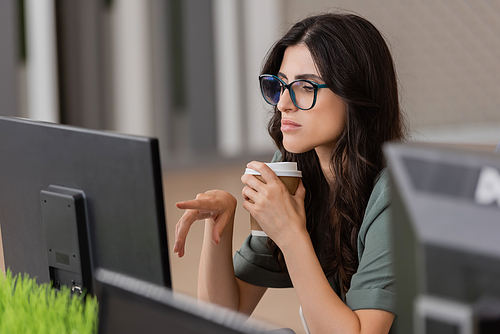 thoughtful businesswoman in eyeglasses holding coffee to go and pointing at computer monitor while working in office