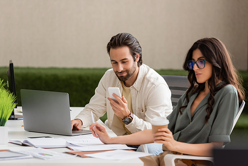 smiling businessman looking at smartphone and using laptop near blurred colleague with takeaway drink