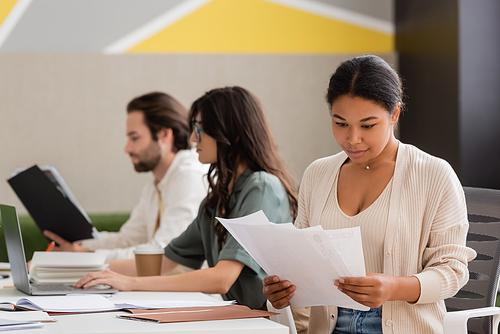 multiracial businesswoman looking at papers near blurred colleagues working with laptop and documents