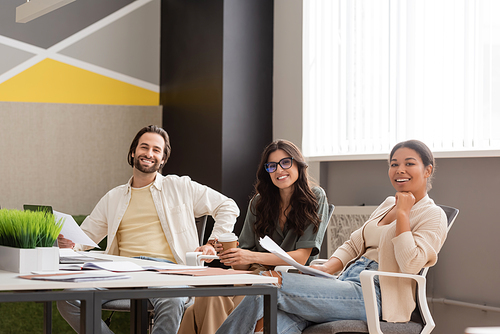 happy multicultural business people looking at camera while sitting near desk with documents in modern office