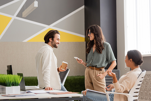 young manager holding paper cup near smiling colleague with smartphone and multiracial businesswoman sitting in office