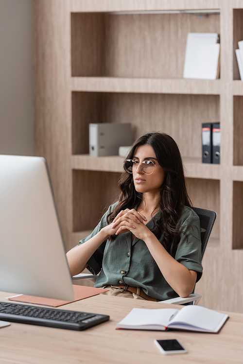 thoughtful manager in eyeglasses looking at computer monitor near empty notebook and mobile phone on work desk