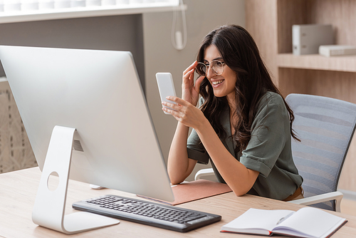 smiling businesswoman adjusting eyeglasses while looking at smartphone near computer and blank notebook