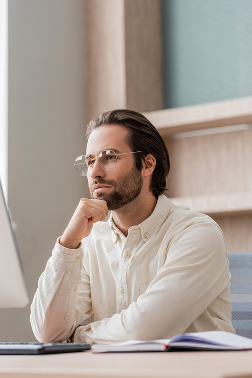 thoughtful businessman in eyeglasses looking at blurred computer monitor in office