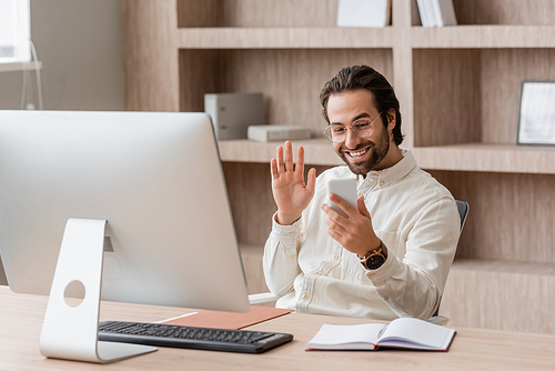 happy bearded businessman in eyeglasses waving hand during video call on smartphone near computer monitor